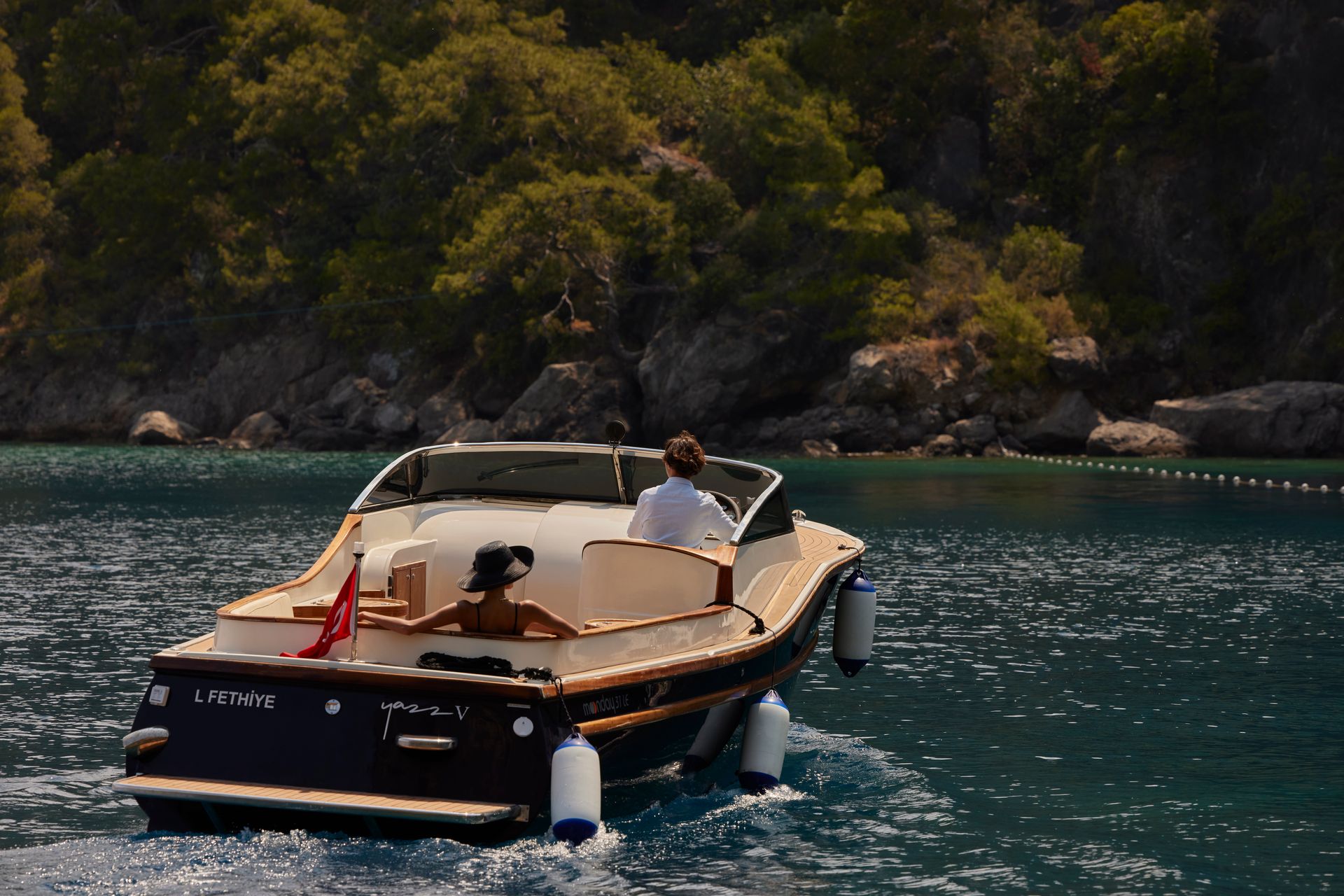 A motorboat carrying two people cruises on blue water toward a tree-covered rocky shoreline.