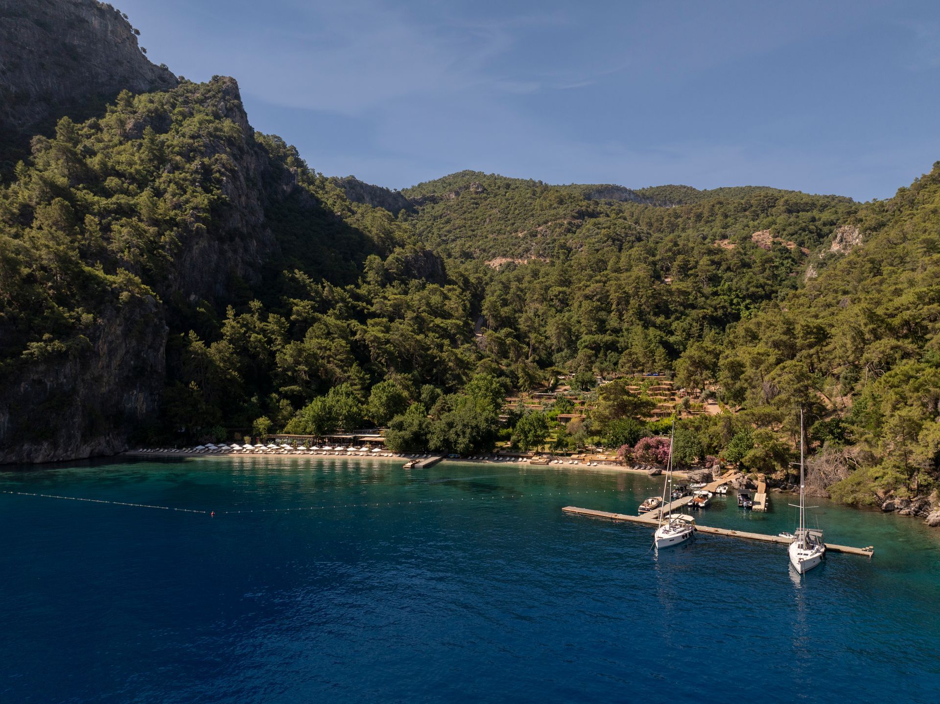 An aerial view of a secluded cove with deep blue water, a small pebble beach, a wooden pier, and dense, forested hills.