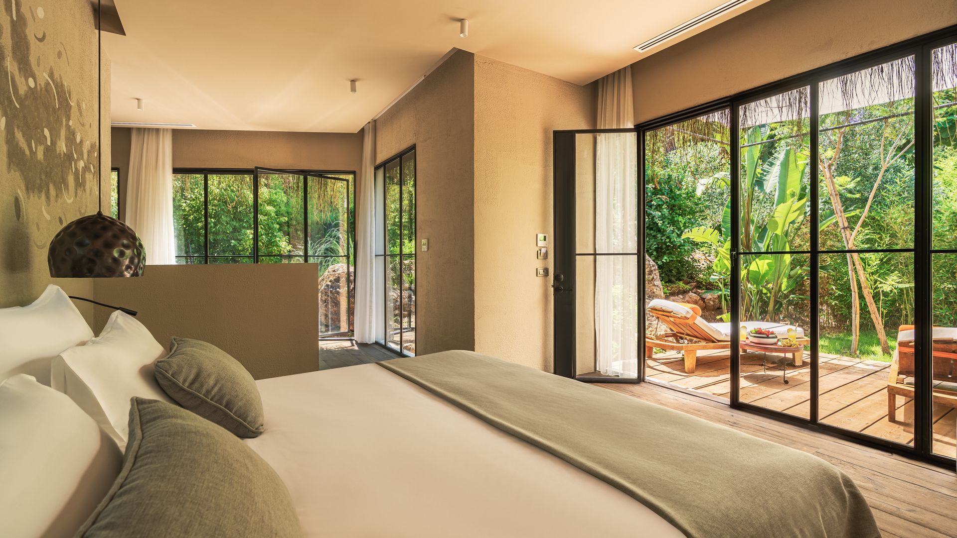 A bedroom with a large bed in the foreground and glass doors opening to a lush, green terrace with lounge chairs.