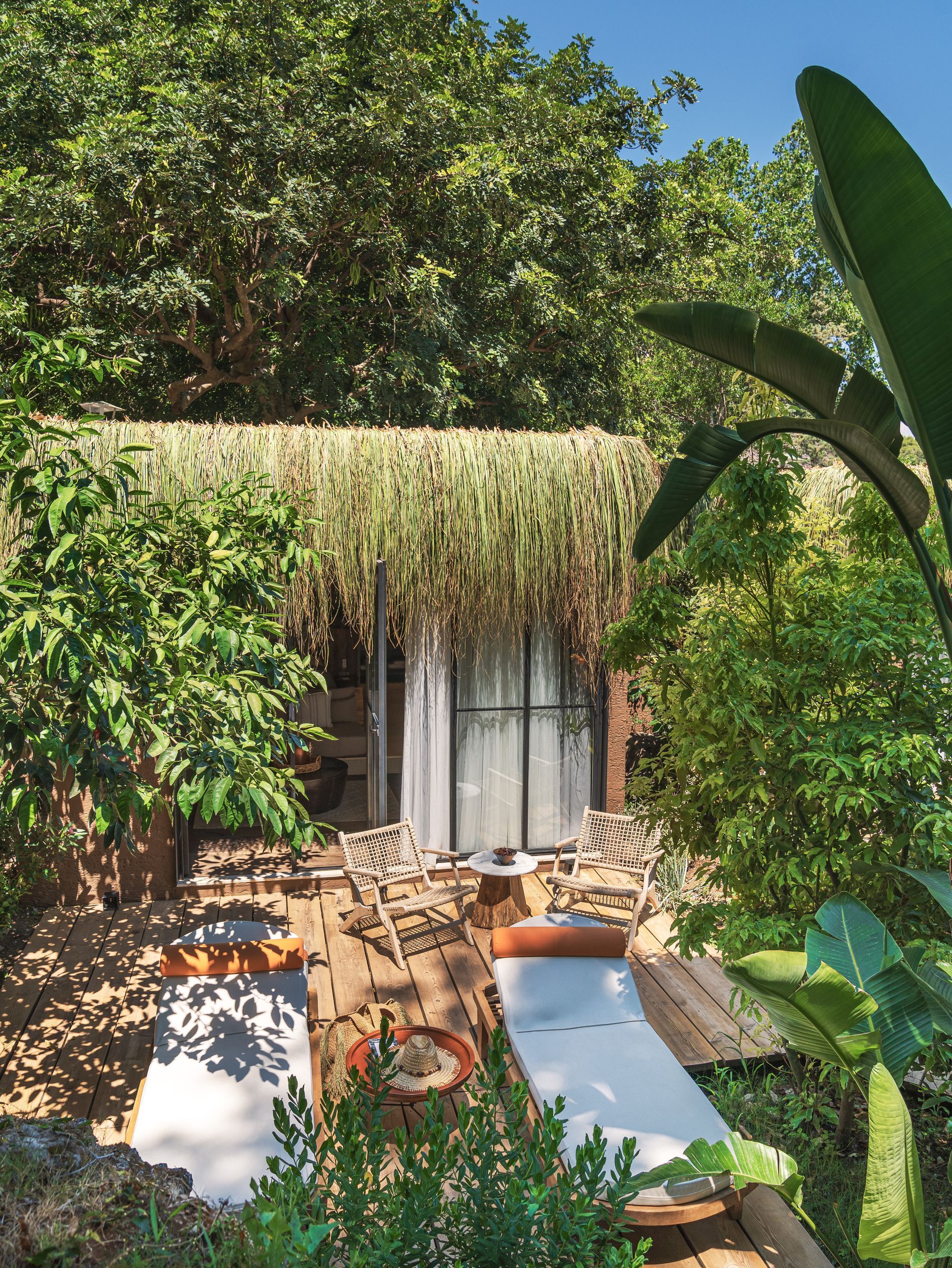 A lush garden patio with two white lounge chairs, small wooden tables, and a small cabana with sheer curtains under trees.