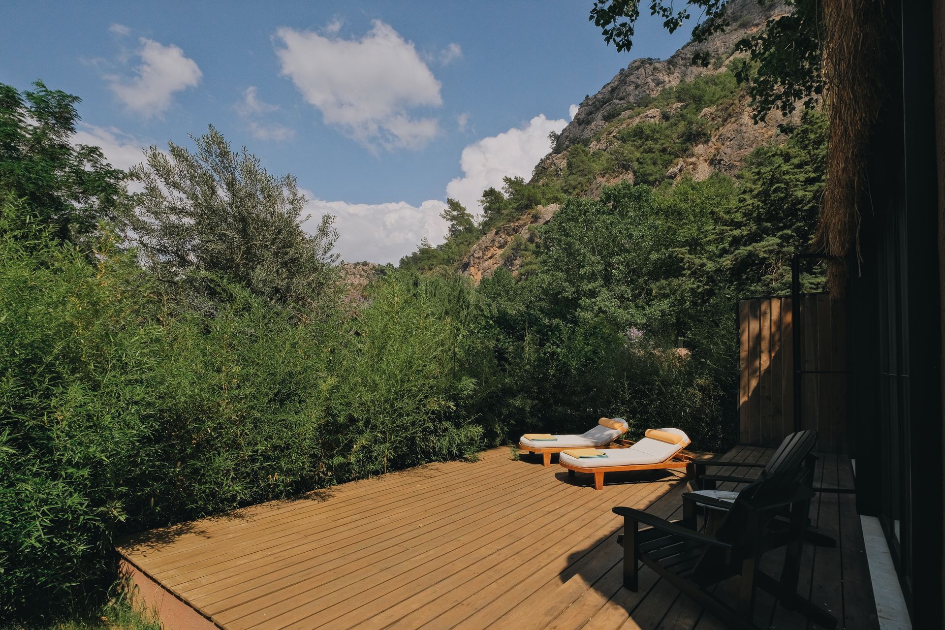 A wooden deck with two lounge chairs and a patio chair overlooking a lush green valley with a mountainside backdrop.