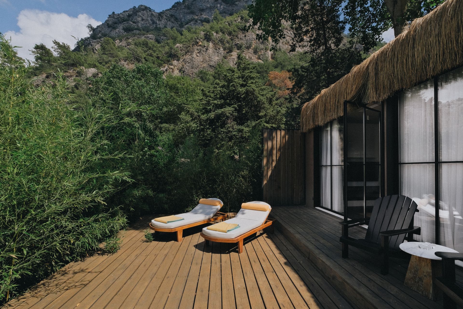 A wooden deck with two lounge chairs and a patio chair outside a modern room with a thatched roof, set against a mountain.