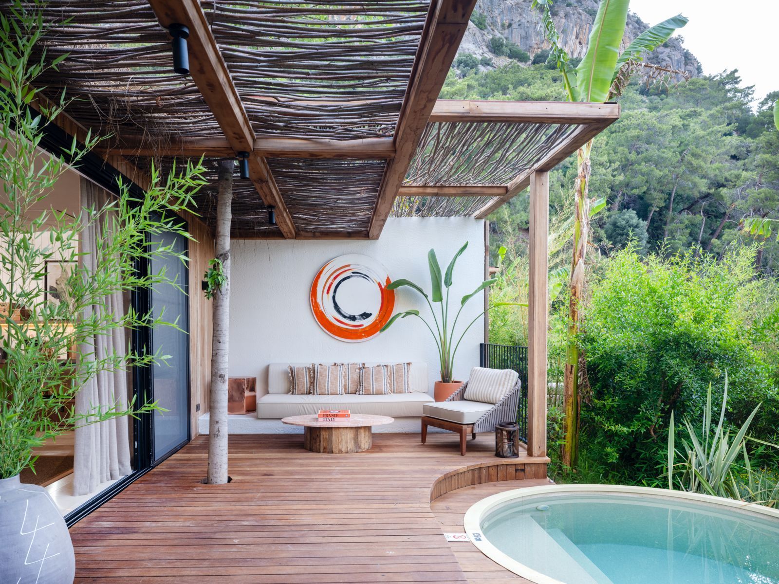 A wooden deck featuring a lounge area with a white sofa, round table, and chair, beside a pool under a rustic canopy.