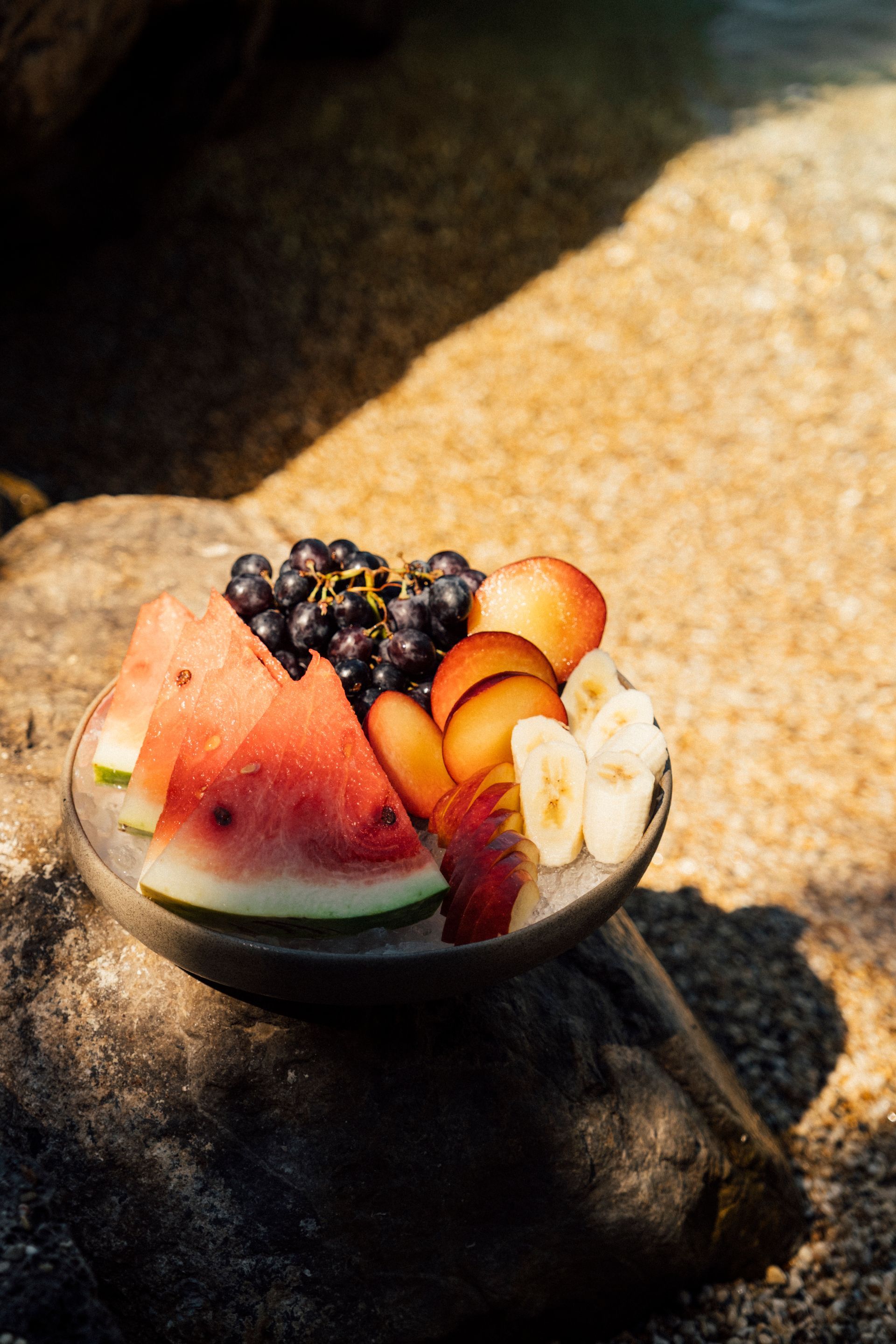 Fruit bowl with watermelon, grapes, orange slices, and pears on a sunlit rock