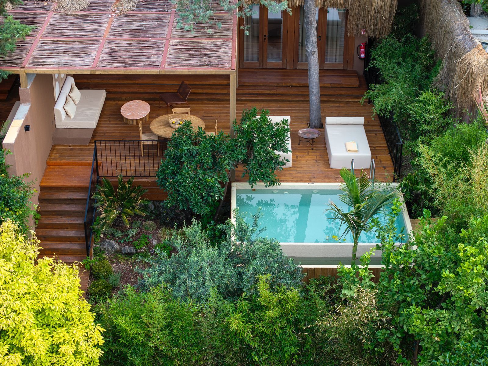 A bird's-eye view of a wooden patio featuring a pool, loungers, outdoor dining, and lush surrounding greenery.