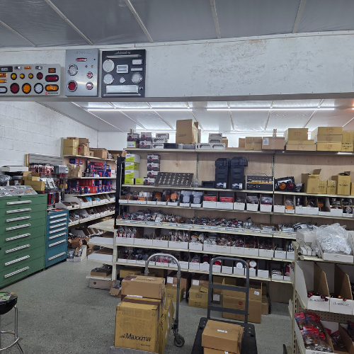 Cluttered warehouse storeroom with shelves of boxes, jars, and supplies under fluorescent lights