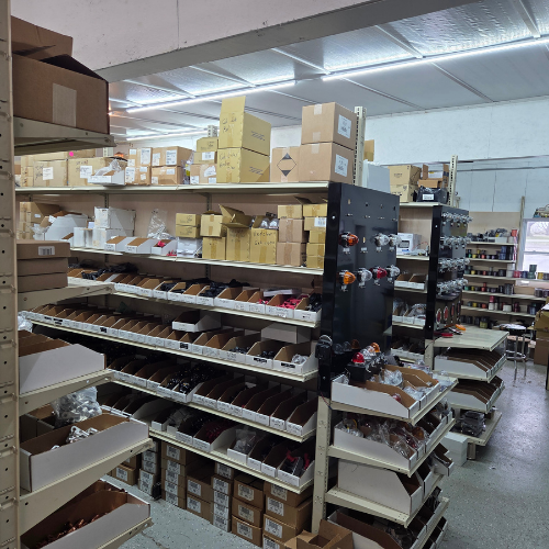 A cluttered stockroom with shelves of boxes, packages, and supplies under bright fluorescent lights.