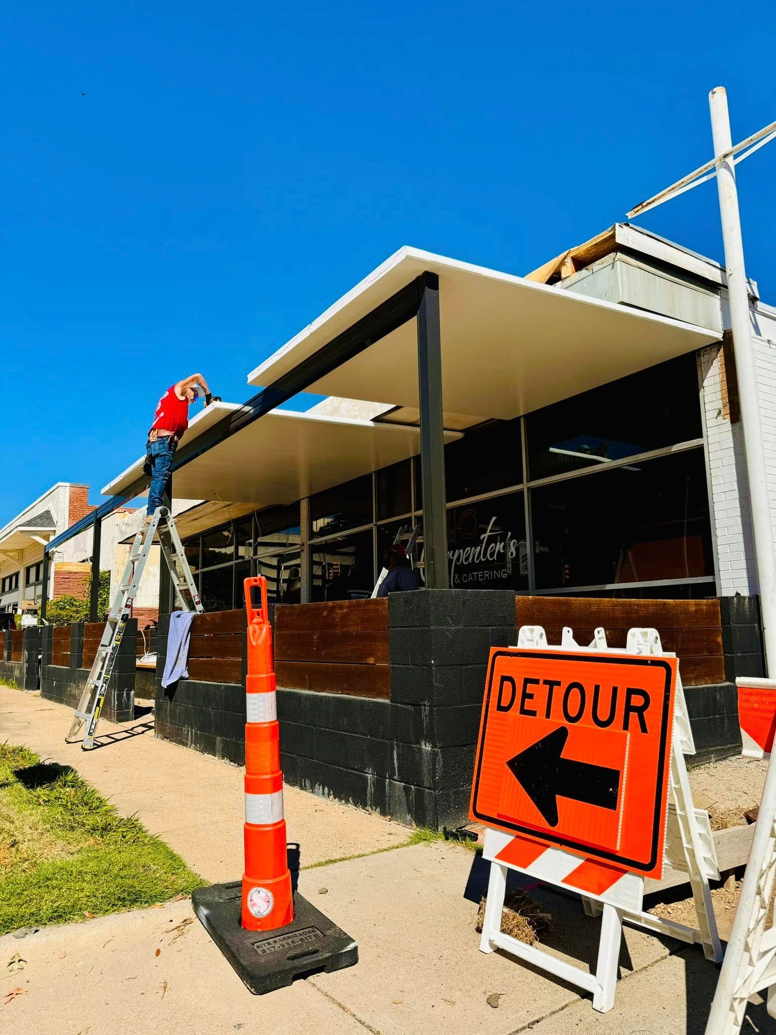 A man on a ladder is working on the roof of a building next to a detour sign.