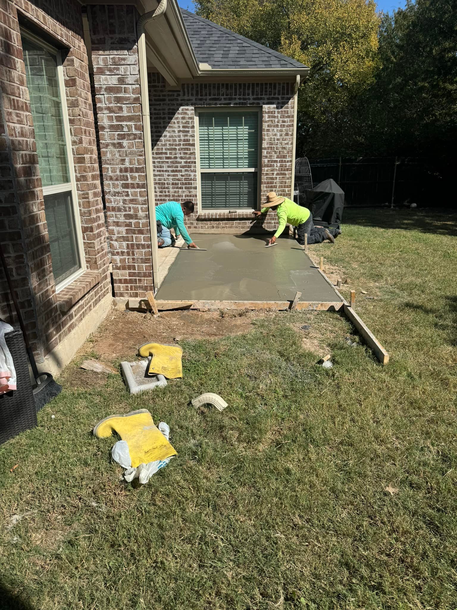 A couple of men are working on a concrete walkway in front of a brick house.