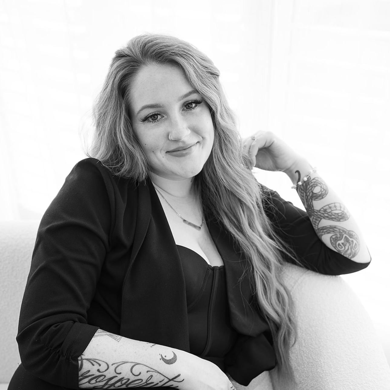 A black and white photo of a woman with long hair and tattoos sitting on a couch.