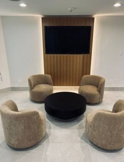 Living room with four beige chairs around a black circular table. A TV is on a wooden wall.