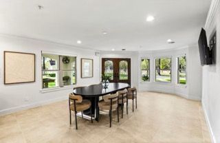 Empty dining room with oval table, chairs, and multiple windows. Pale floor, white walls, and a mounted TV.