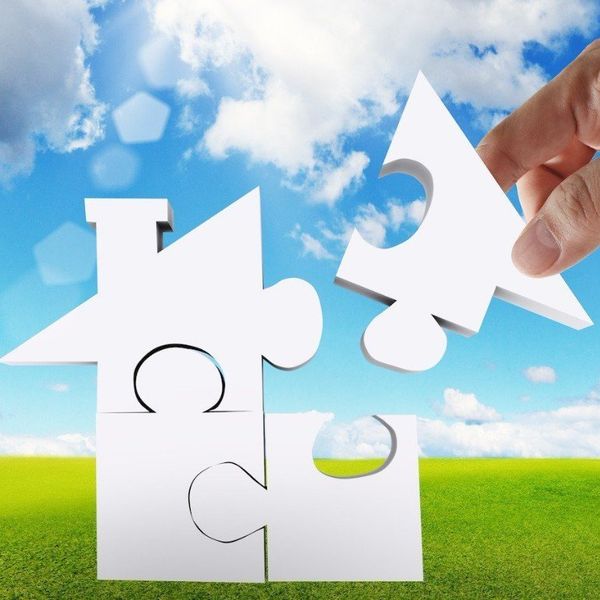Illustration of a puzzle in the shape of a house with a hand placing the final piece. Sky and grass background.