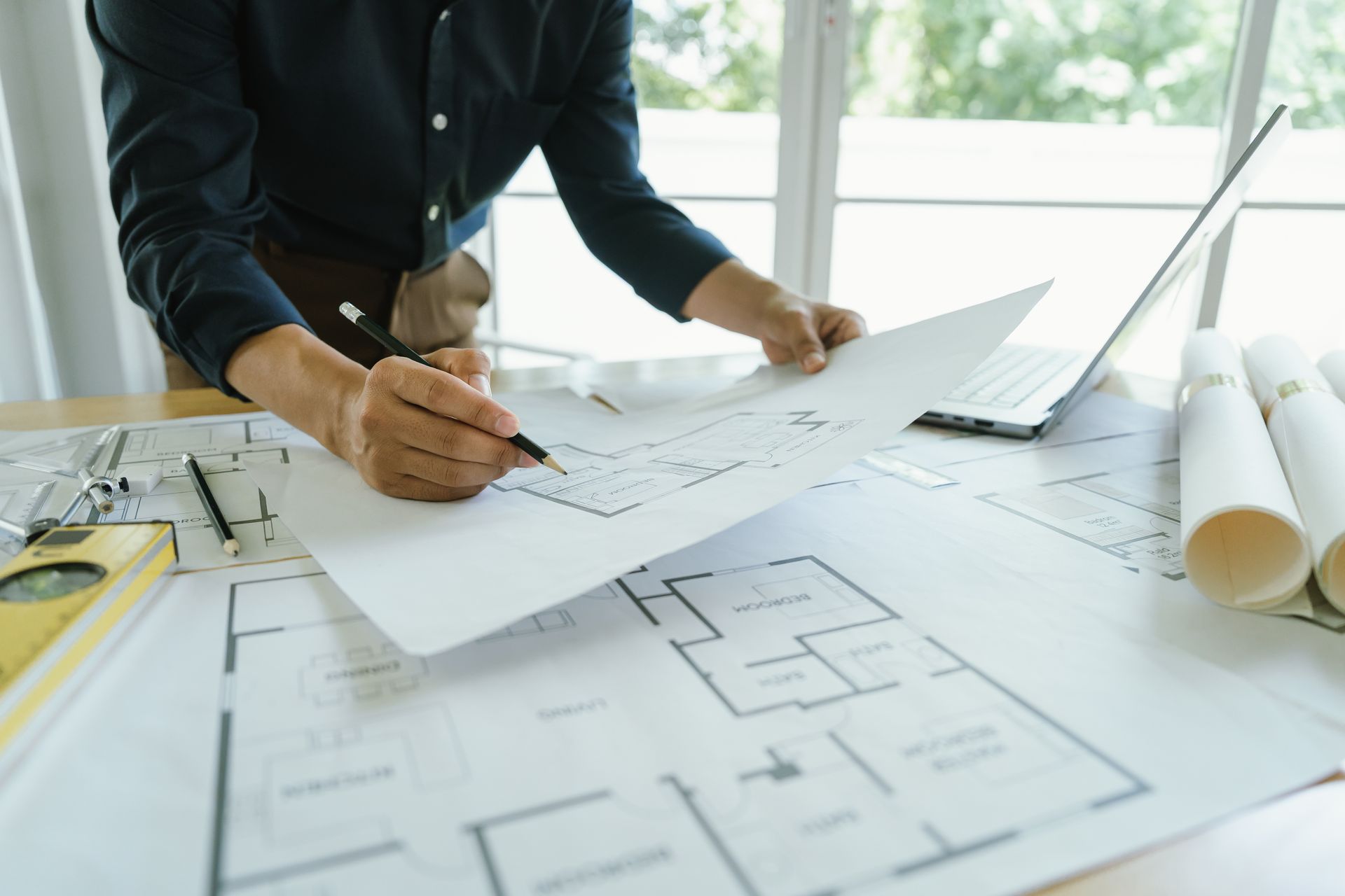 A person in a dark shirt uses a pencil to review architectural floor plans on a desk next to a laptop and rolled blueprints.