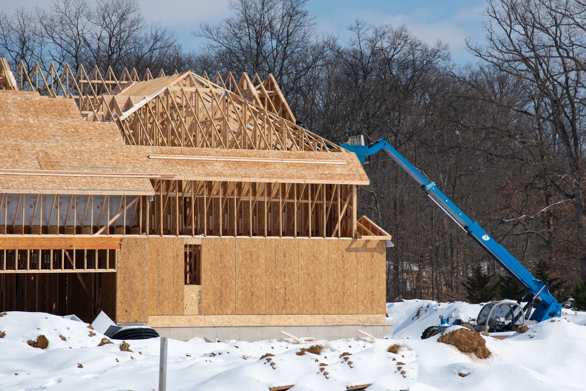 Construction of a house with a blue boom lift, snow on the ground and trees in the background.