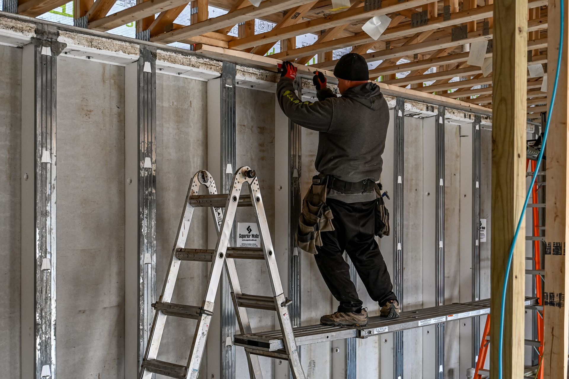 A construction worker on a ladder, installing boards to a metal frame wall. Indoors, daylight.