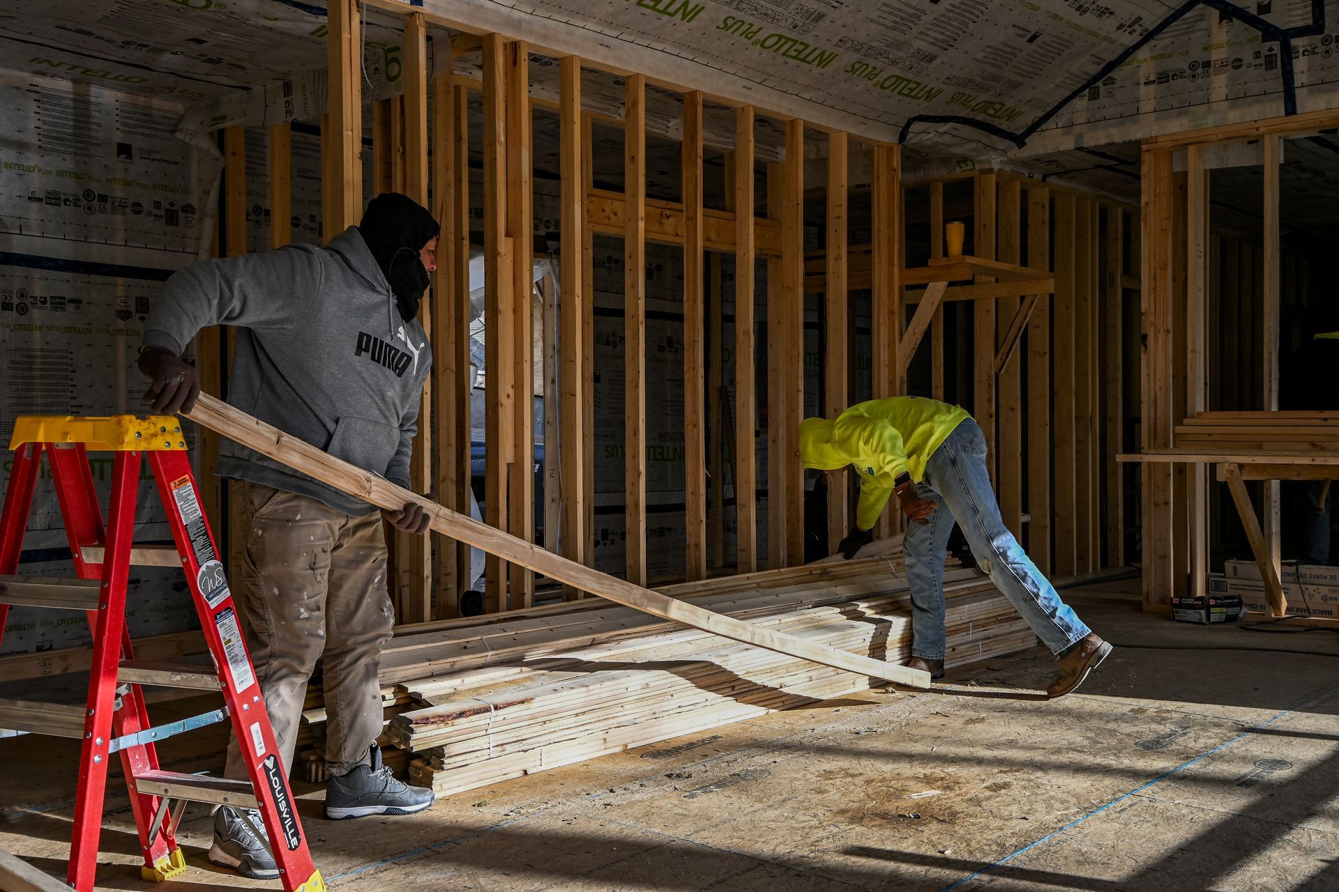Two construction workers framing walls inside a building under construction. One holds lumber, the other bends down.