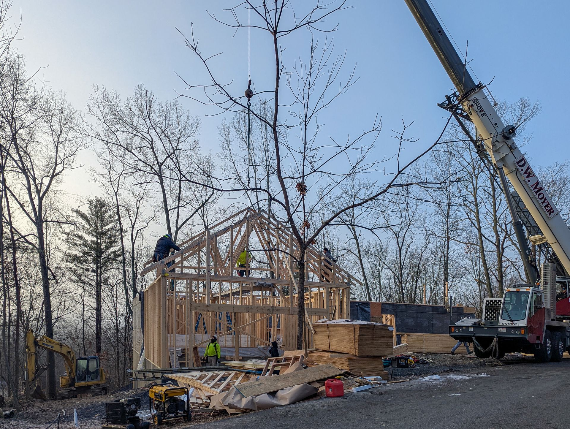 Construction workers assemble a wooden frame building using a crane in a wooded area.