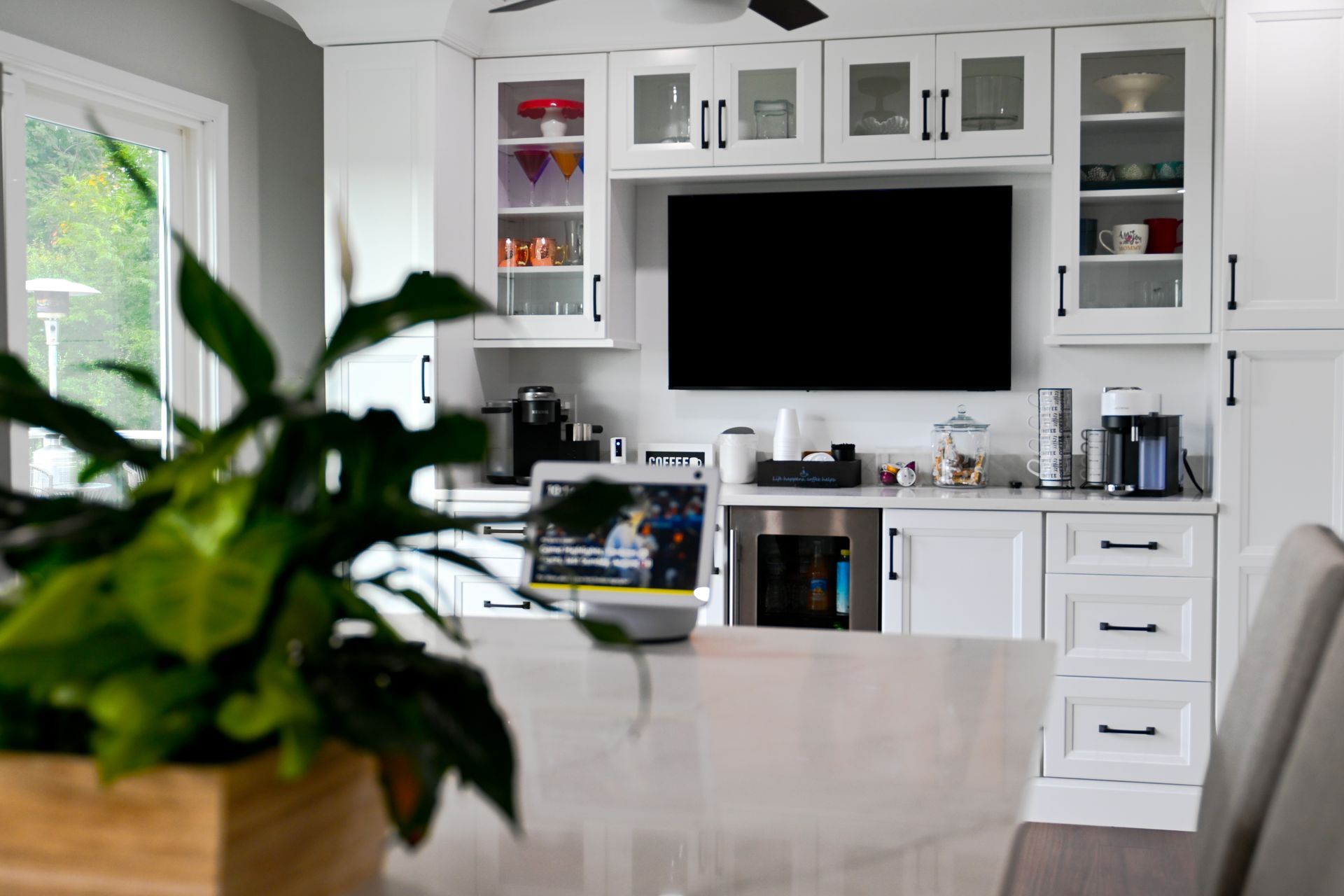 Kitchen with white cabinets, built-in TV, and countertop appliances. Plant in foreground, window on left.