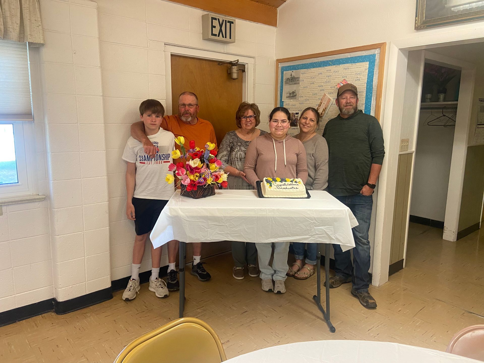 Five people stand behind a table with a colorful bouquet and cake in a hallway near an exit sign.