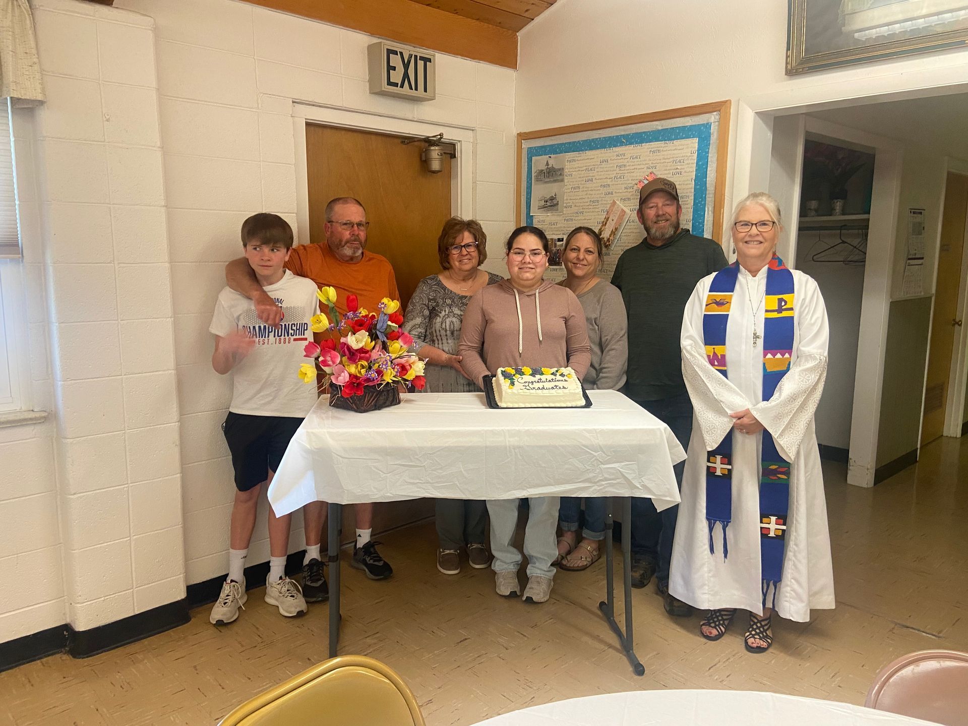 Group posing indoors beside a table with flowers; a priest stands at right in white vestments.