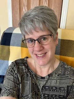 Woman with short gray hair, glasses, smiling, sitting. Wearing a patterned top, plaid blanket behind.