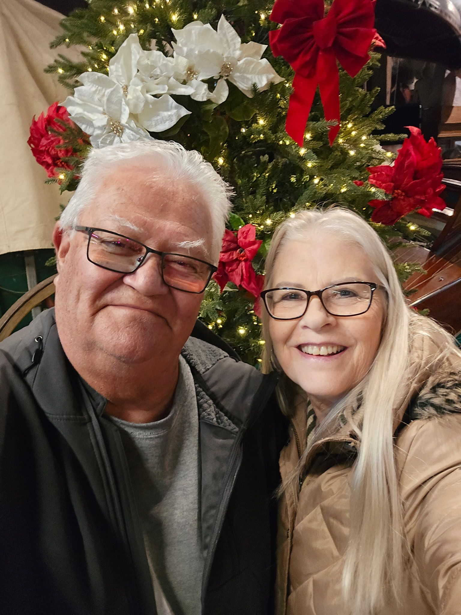 A smiling couple stands together in front of a Christmas tree decorated with white poinsettias and a large red bow.