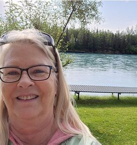 Woman with glasses smiles outdoors near a river and small dock.