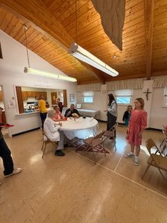 People gathered in a bright hall with a wood ceiling, some seated at round tables.