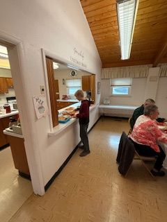 Person serving food from a kitchen counter; other people sit at a table in a room with a wooden ceiling.