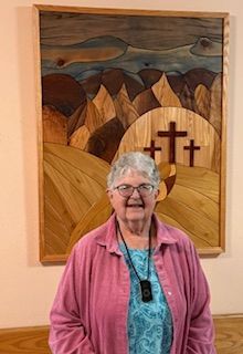 Woman in pink shirt standing in front of wood art of a landscape and three crosses.