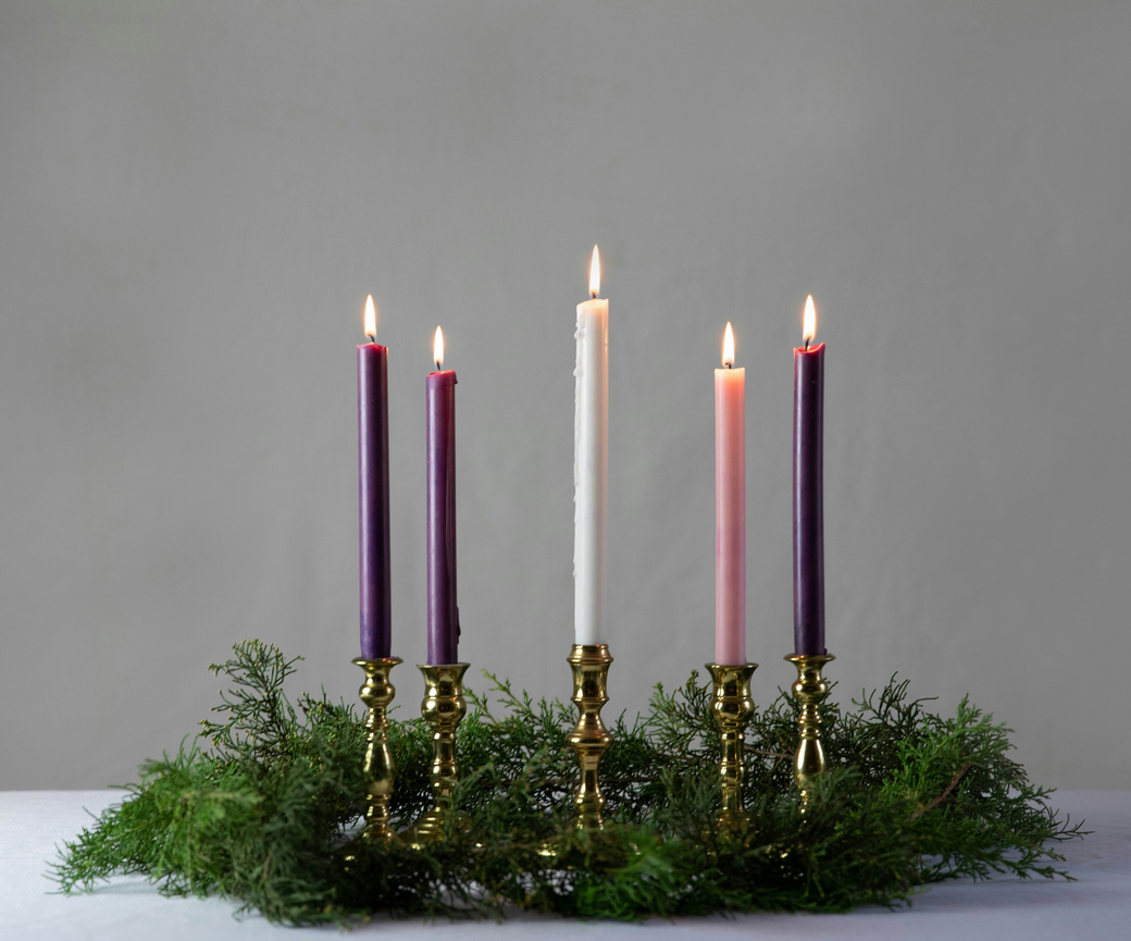 Lit Advent wreath with four purple candles, one pink, and one white, surrounded by greenery.