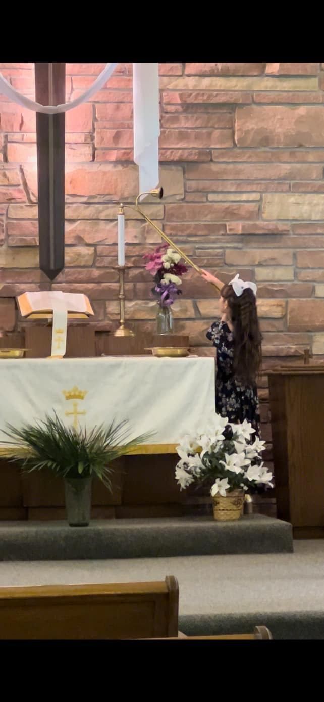 Person lighting a candle with flowers in a church. The person has long hair and is wearing a dark floral dress.