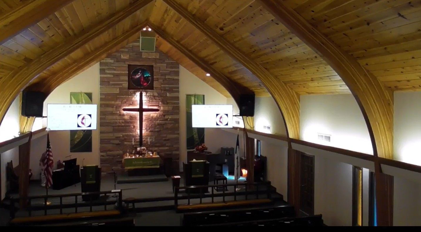 Interior of a church with a wooden ceiling, cross, two screens and pews.