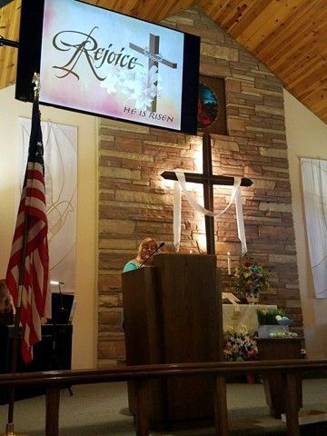 Woman speaking at a church podium with a cross and screen displaying 