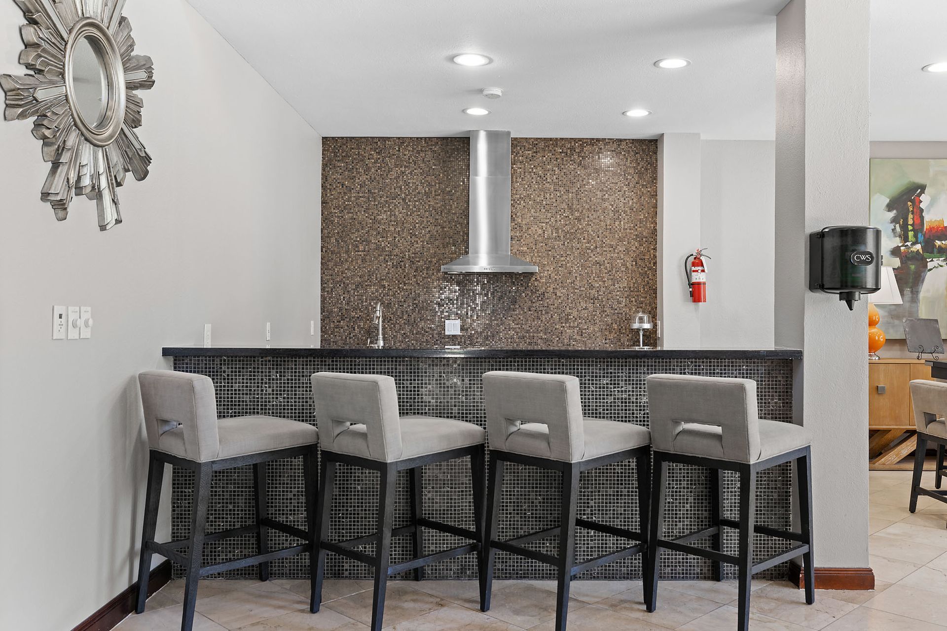 Communal kitchenette/bar area with mosaic tile backsplash and four gray upholstered bar stools.