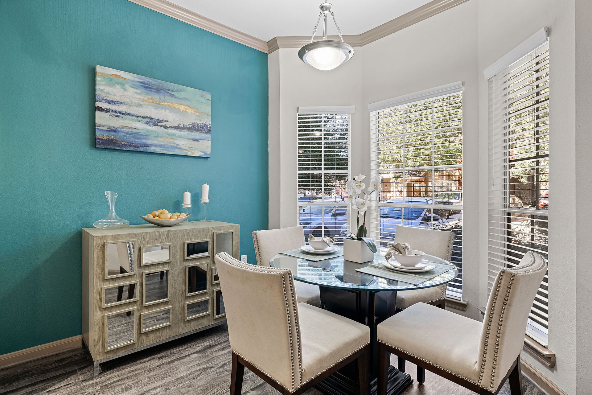 Dining nook in an apartment with teal accent wall, glass table, beige chairs, and large windows with blinds.