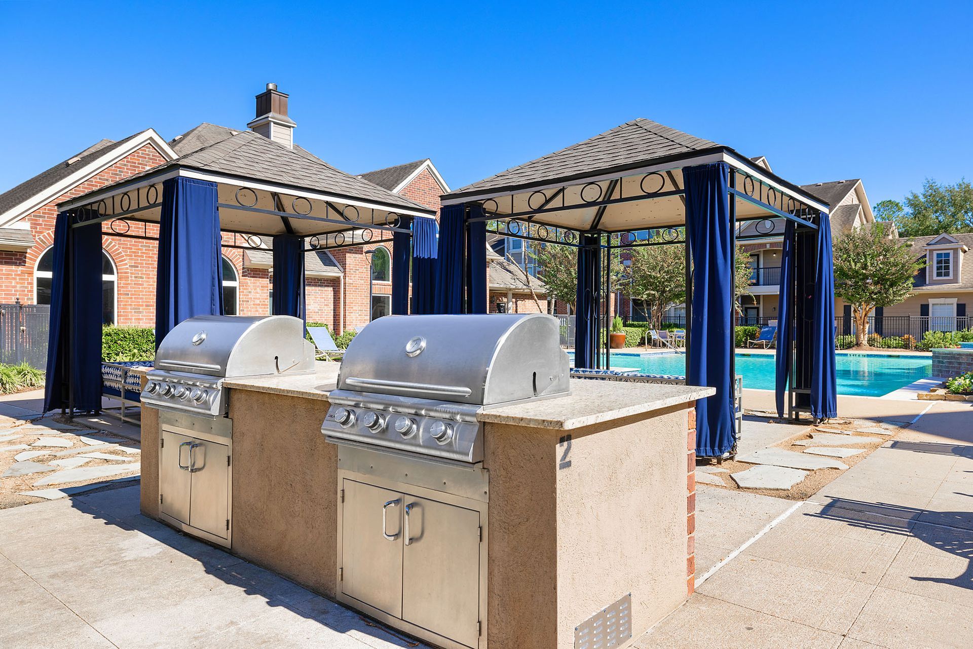 Outdoor communal barbecue grills under blue canopies beside a swimming pool.