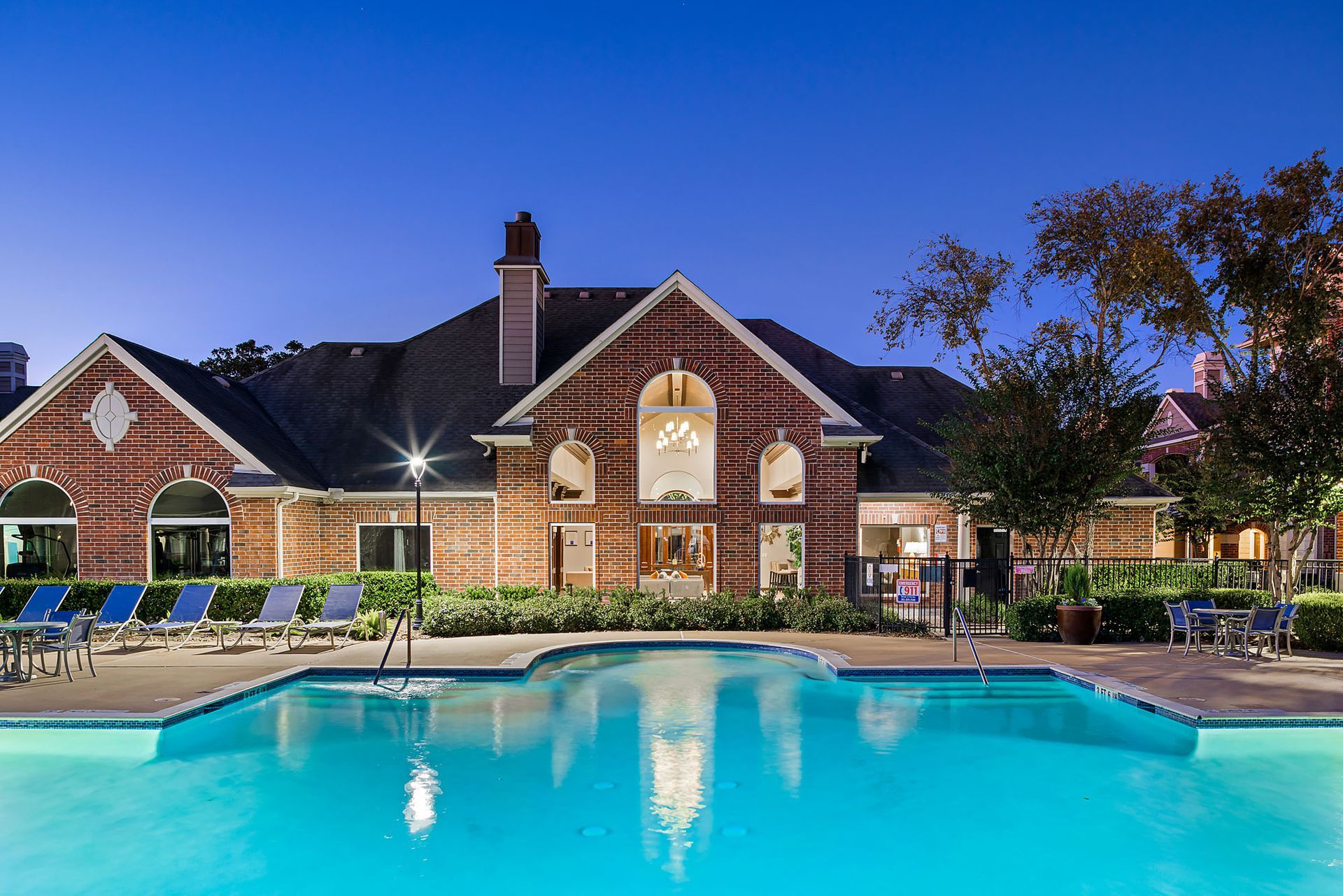 Exterior view of a brick apartment community with a lit pool and lounge chairs.