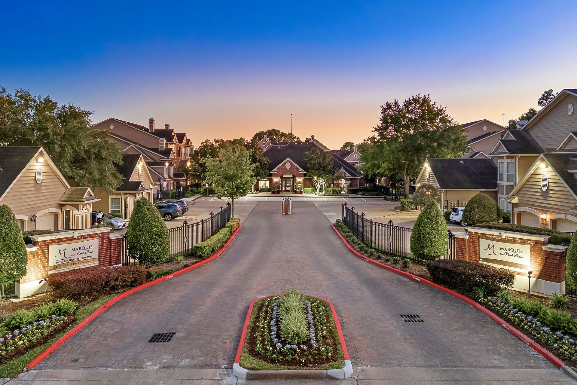 A gated apartment community entrance with brick signage, trees, and landscaped medians at dusk.