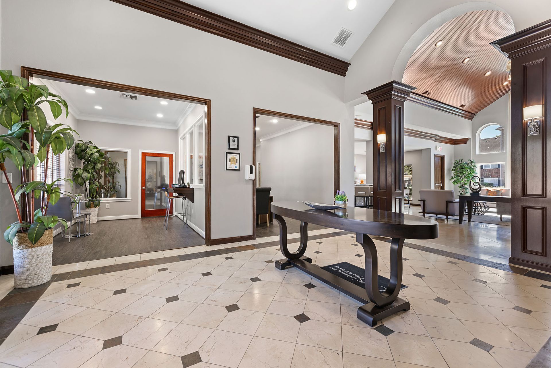Lobby area with tiled floor, dark wood trim, arches, plants, and a reception desk.