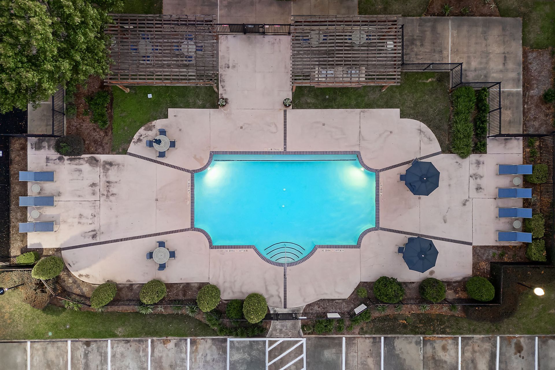 Aerial view of a rectangular blue pool surrounded by a concrete deck, lounge chairs and umbrellas