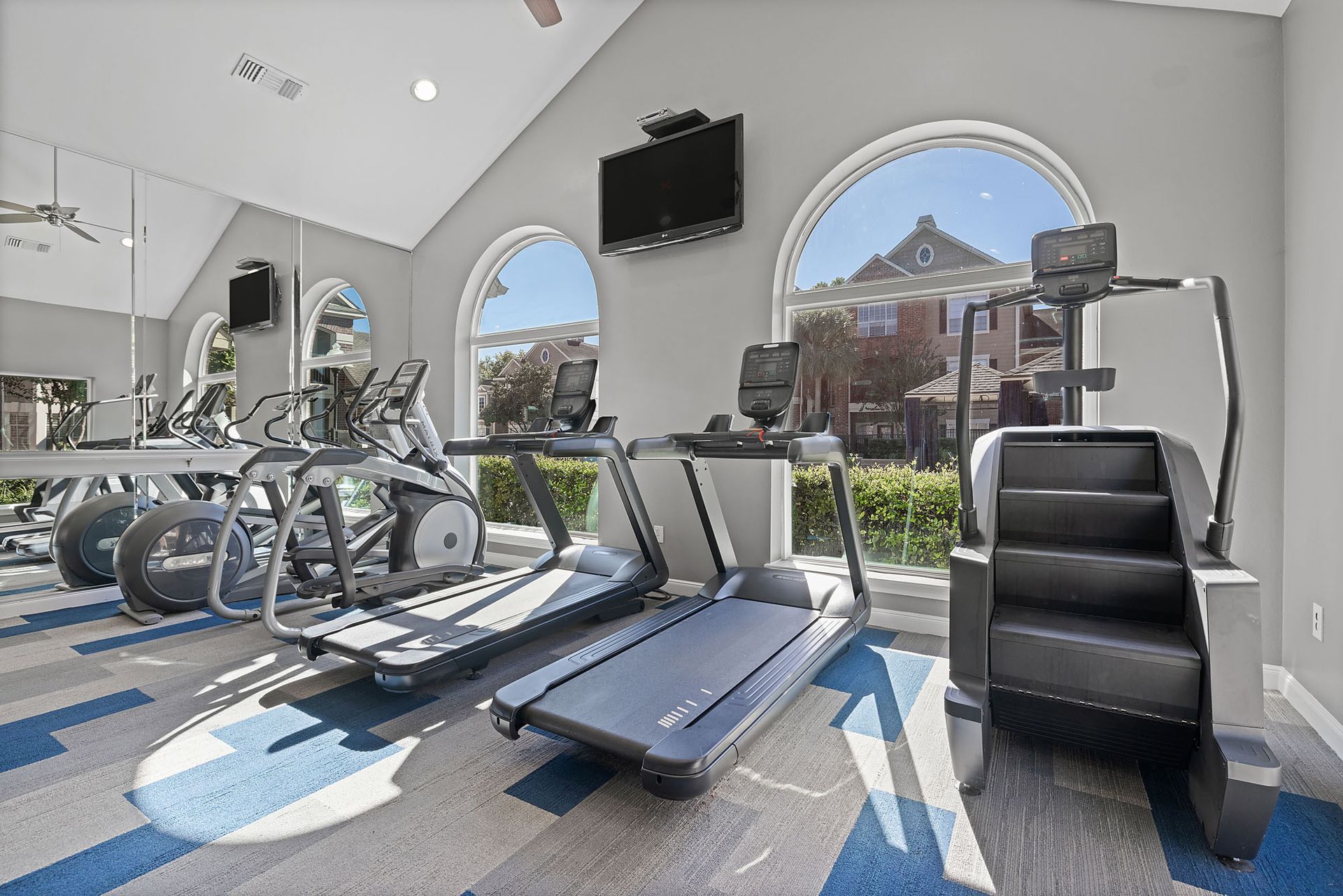 Indoor community gym with treadmills and ellipticals, a wall-mounted TV, and large arched windows.