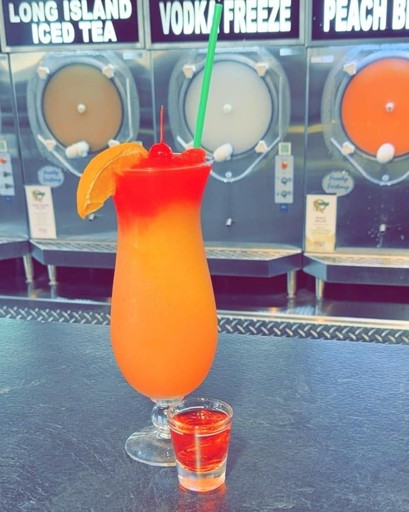 Tropical cocktail with orange and cherry garnish, next to a shot glass on a bar, with drink machines in the background.