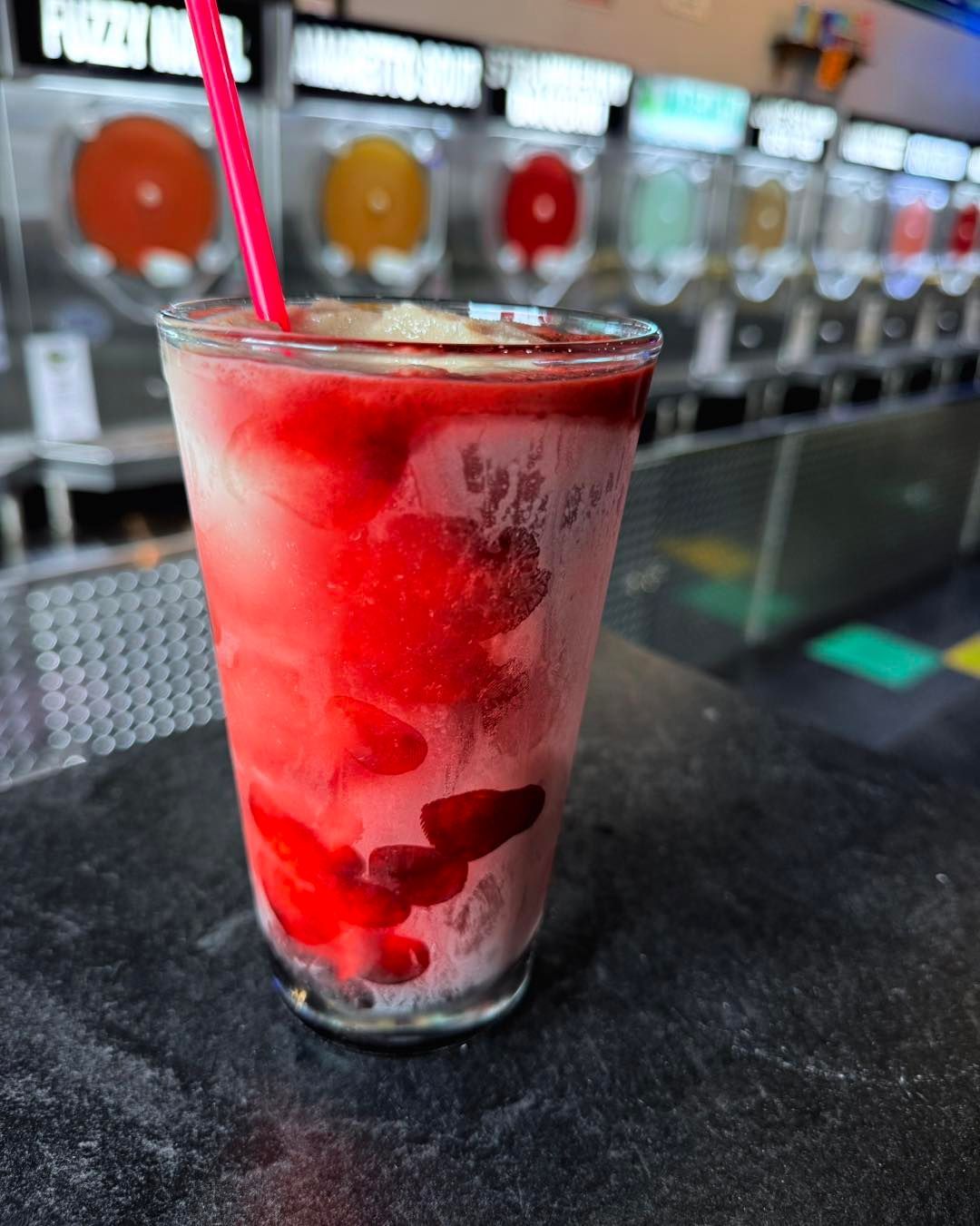 Glass of red and white frozen drink with a pink straw. Bar setting, multiple drink dispensers in the background.