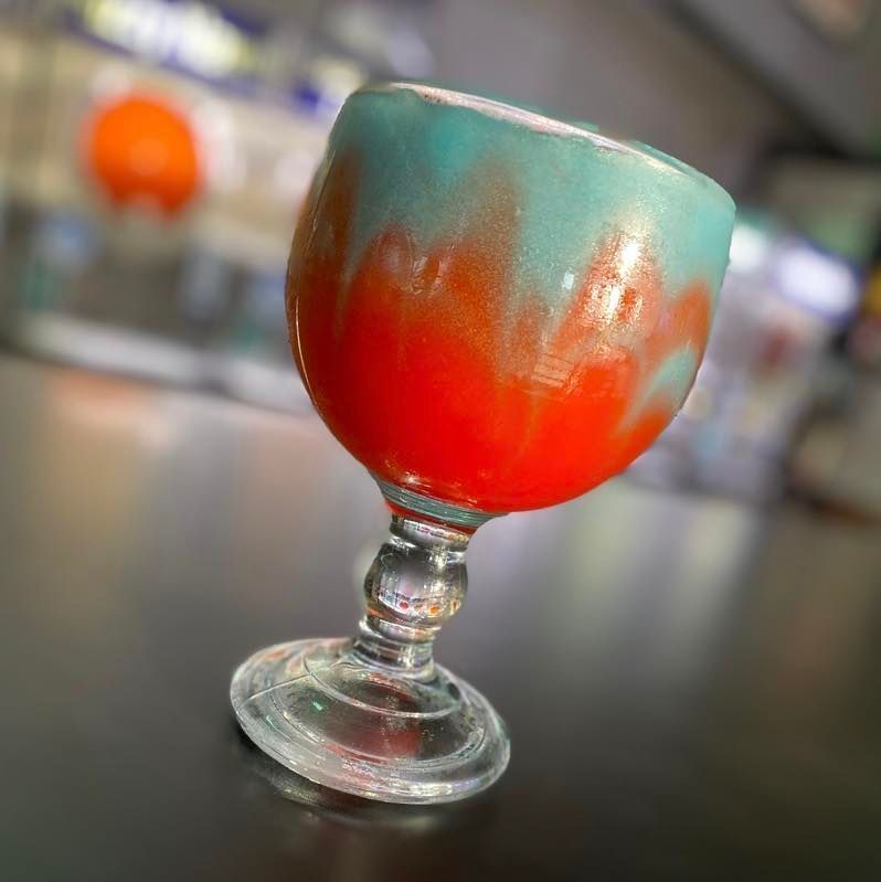 Glass of red and teal liquid on a black surface; orange out-of-focus background.