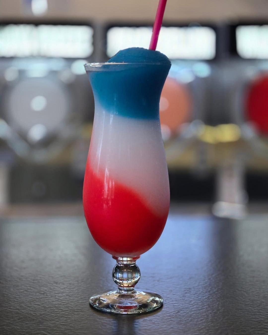 Red, white, and blue layered frozen drink in a stemmed glass with a pink straw, on a dark bar.