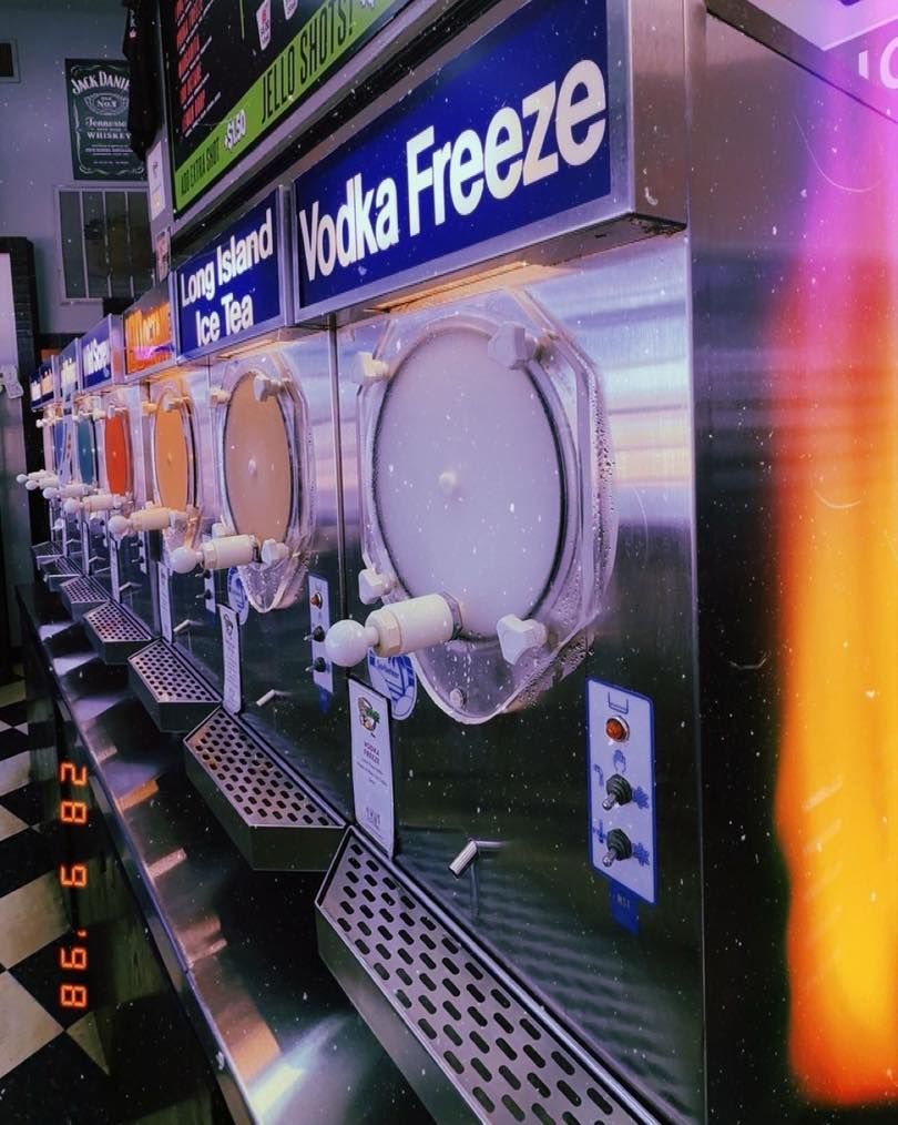 Slushy machines dispensing frozen drinks. Vodka Freeze label above the central machine, other flavors visible. Checkerboard floor.