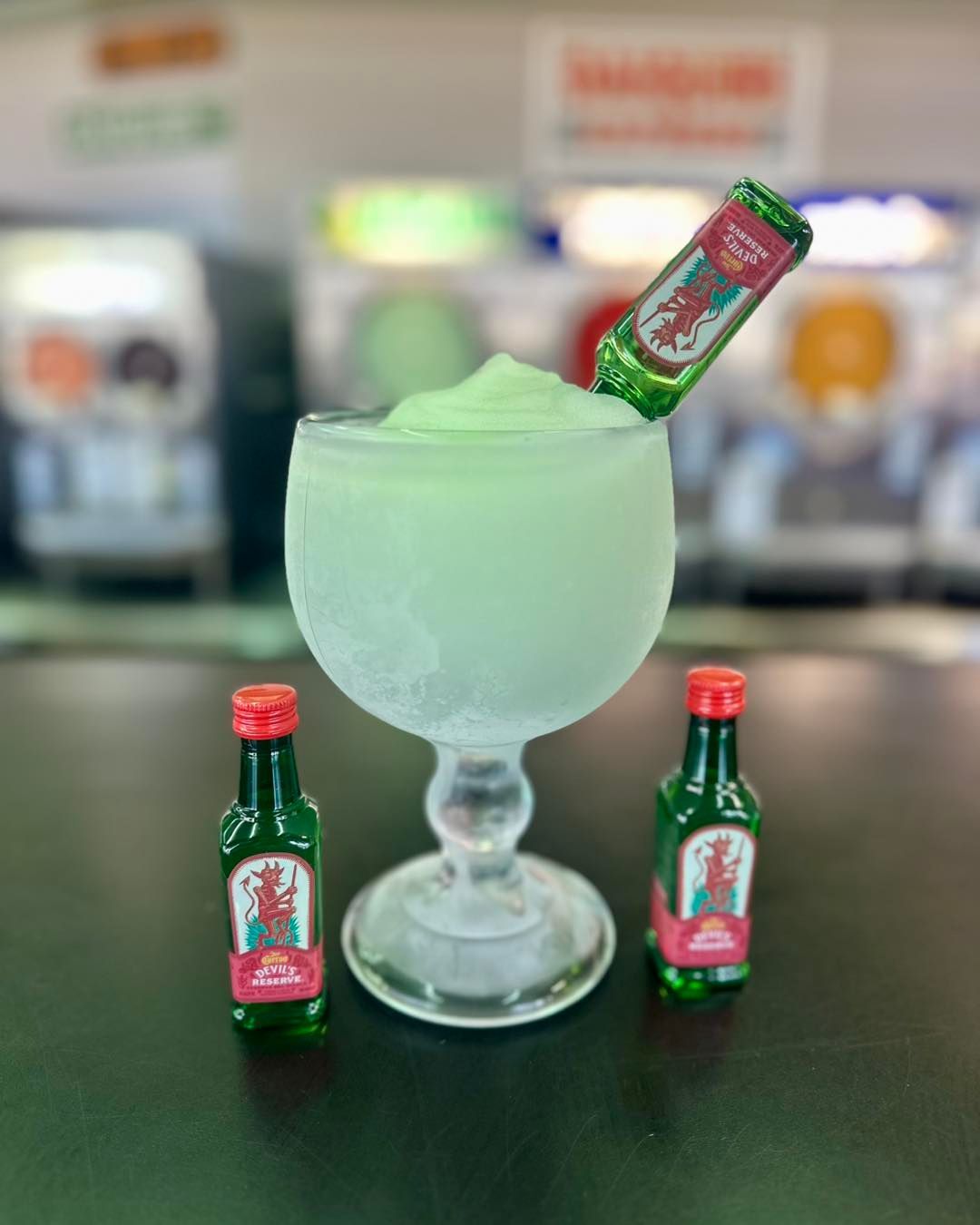 Frozen green cocktail in a stemmed glass with mini liquor bottles on a bar.
