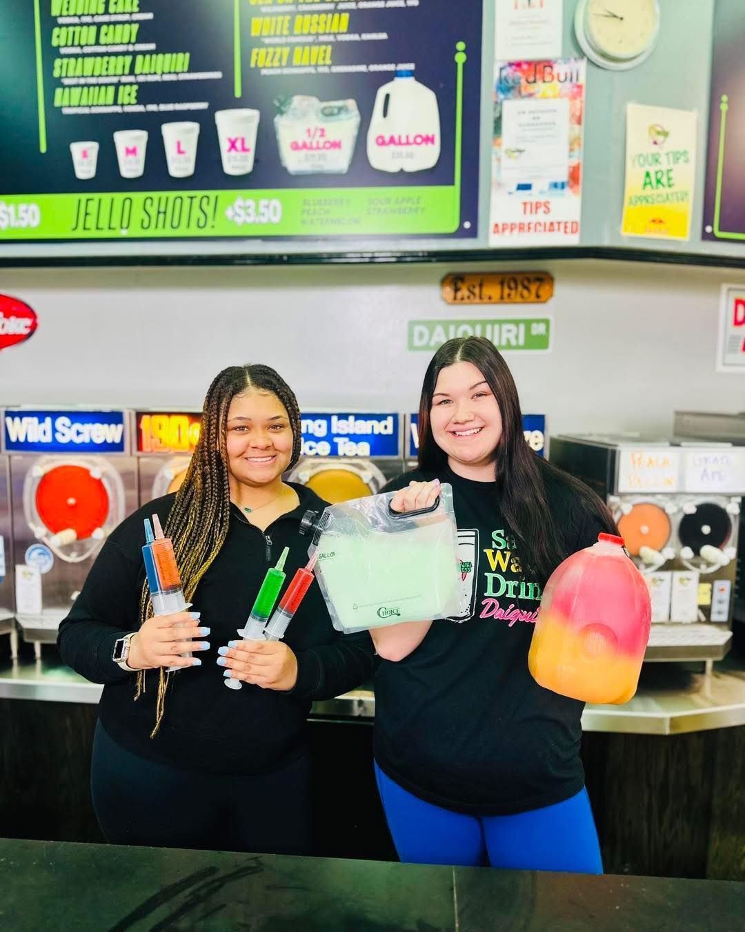 Two women smiling, holding slushy drinks and jello shots behind a counter.
